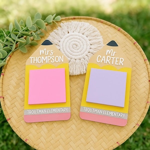 May include: Two pencil-shaped memo boards with yellow bodies, pink and gray erasers, and black tips. One board reads "Mrs. Thompson" and the other "Mr. Carter." Both have a sticky note pad and the text "Troutman Elementary."