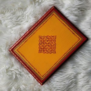 May include: A rectangular, hand-painted wooden box with a vibrant yellow surface. The box features a red border with gold accents and a central, intricate red design. The box is set against a fluffy, white background.