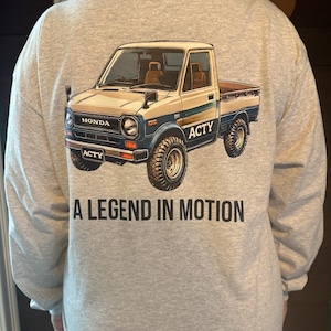 May include: A gray long-sleeve shirt with a graphic of a white Honda Acty pickup truck. The truck has blue accents and the text "A LEGEND IN MOTION" is printed below the truck.