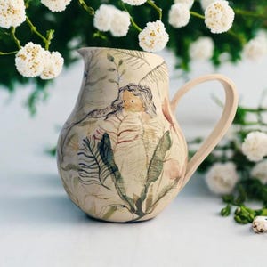 May include: A ceramic pitcher with a cream-colored base, featuring a hand-painted design of a woman surrounded by botanical elements. The pitcher has a curved handle and is adorned with green, brown, and pink foliage. The overall aesthetic is artistic and nature-inspired.