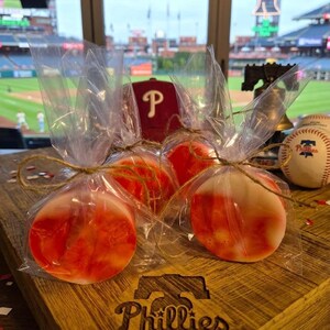 May include: Three round, red and white treats wrapped in clear cellophane and tied with twine, presented on a wooden board with a Phillies logo. A baseball and a Phillies cap are in the background.