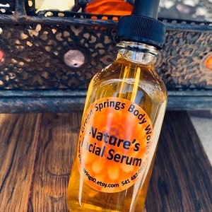 May include: A clear glass bottle with a black dropper top filled with a yellow liquid. The bottle has a round orange label that reads "Green Springs Body Works - Nature's Facial Serum - greensprings0.etsy.com 541 482-0857".