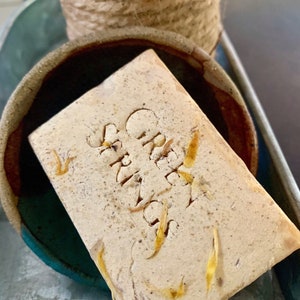 May include: A rectangular bar of soap with the word "Greek" carved into it, resting in a ceramic bowl. The soap is light brown with yellow accents. A woven rope container is in the background.