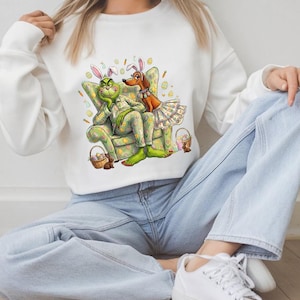 May include: White sweatshirt featuring a cartoon Grinch and a dog wearing bunny ears and Easter attire. The Grinch is seated in a patterned armchair, with Easter baskets nearby. The design is colorful and festive, perfect for Easter.