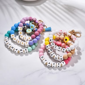 Könnte beinhalten: Bunte Schlüsselanhänger mit Namen, die in weißen Buchstabenperlen geschrieben sind. Die Schlüsselanhänger haben eine Vielzahl von Farben, darunter Rosa, Blau, Gelb und Weiß. Einige Schlüsselanhänger haben dekorative Perlen und Quasten.