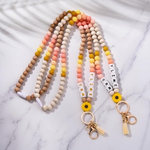 May include: Beaded lanyards in brown, yellow, and pink. Each lanyard has a name spelled out with white letter beads. Gold-coloured key rings and sunflower charms are attached to the lanyards.