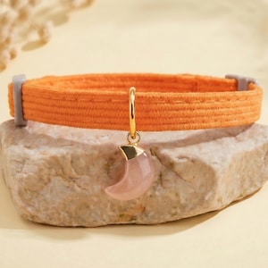 May include: An orange fabric collar featuring a gold-toned metal ring and a smooth, pale pink crescent moon-shaped charm. The collar includes grey plastic hardware components on each side, providing a secure closure for the adjustable band.