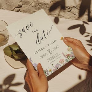 May include: A cream-colored "Save the Date" card with floral accents is held by hands. The card features elegant script and the names "Elisha + Daniel" along with the date "June 25 2026". A glass, plate, and cup are nearby.