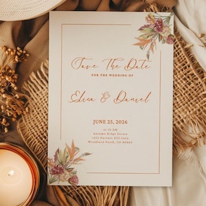 May include: A wedding invitation with the text "Save The Date" and the names "Elisa & Daniel" is displayed. The invitation is decorated with floral accents and the date June 25, 2026. A straw hat, candle, and dried plants are arranged around the invitation.