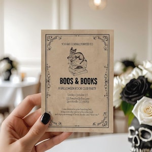 This is a lifestyle image of dinning room - white walls, black skull accents in the background - in the foreground is a womens hand holding a vintage inspired white card. The card is a Halloween invitation for a Boos and books - or a Halloween book club invite. The card has an illustrated black cat and books. This card is vintage inspired.