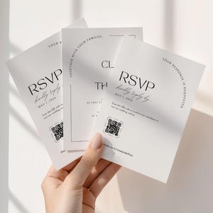 This is a lifestyle image of a womens hand holding three of the wedding invite templates up against a plain white wall. The invites are plain white with bold, classic inspired fonts.