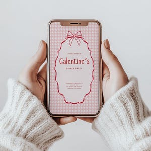 May include: A smartphone screen shows a pink Galentine's Dinner Party invitation. The invitation has a bow and an oval frame with text. The background has a repeating pattern. The phone is held by hands wearing a cream-coloured jumper.