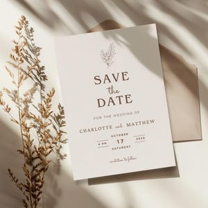 May include: A cream-coloured wedding invitation with the text "Save the Date" in a brown serif font. The invitation includes the names Charlotte and Matthew, the date October 17, 2026, and the time 4 PM. A dried plant and envelope are also visible.