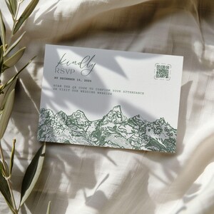Peut inclure: Carte RSVP blanche avec texte vert et illustration de montagnes. La carte indique "Kindly RSVP" avec une date limite au 15 décembre 2025. Comprend un code QR et des instructions pour confirmer la présence ou visiter le site web du mariage.