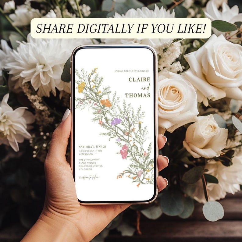 This is a lifestyle mockup photo of a manicured hand holding a phone in front of background of white roses. In the center of the phone screen is an wildflower wedding invite.