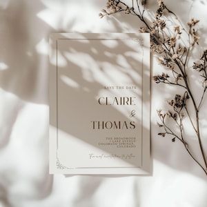 May include: A cream-colored wedding invitation with the text "SAVE THE DATE" and the names "CLAIRE & THOMAS" in a serif font. The invitation also includes the wedding location and the phrase "Formal invitation to follow."