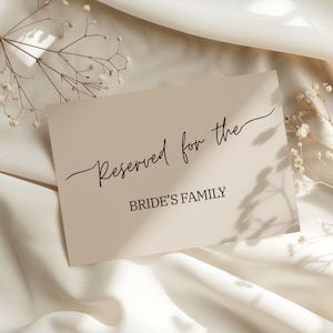 May include: A beige card with the text "Reserved for the BRIDE'S FAMILY" in black script and sans-serif font. The card is placed on a white, textured fabric, with delicate white flowers and shadows in the background.