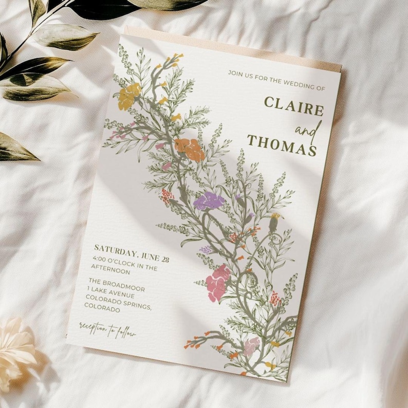 This is a lifestyle mockup photo of a wedding invitation on a linen table. There are a dried olive branch, and dried willow branch on the table, there are shadows that overlay the invitation. The invite has hand illustrated floral sprigs.