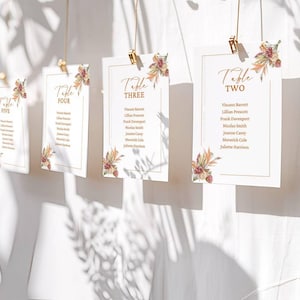 May include: Elegant wedding table cards with gold lettering and floral accents. Each card is rectangular and features the word "Table" followed by a number, along with guest names. The cards are suspended by gold clips.