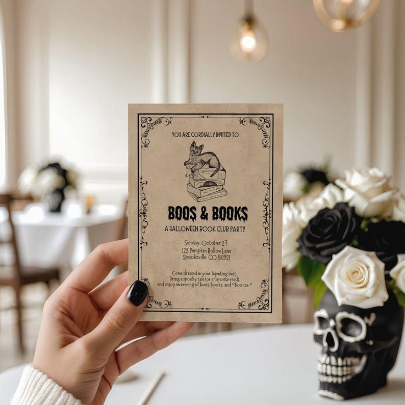 This is a lifestyle image of dinning room - white walls, black skull accents in the background - in the foreground is a womens hand holding a vintage inspired white card. The card is a Halloween invitation for a Boos and books - or a Halloween book club invite. The card has an illustrated black cat and books. This card is vintage inspired.