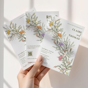 This is a lifestyle image of a womens hand holding three of the wedding invite templates up against a plain white wall. The invites are in a summery green, orange, yellows, purple, pinks against a bright white card.