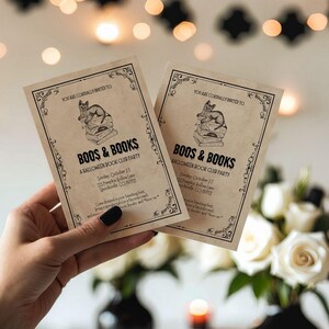 This is a lifestyle image of dinning room - white walls, black skull accents in the background - in the foreground is a womens hand holding  two vintage inspired cards. The card are for a Halloween invitation - for a Halloween themed book club.