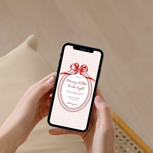 This is a lifestyle image mockup of a manicured pair of hands holding a phone. On the phone screen is a Christmas party invitation - this one is white and red.