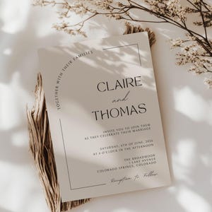 Op de afbeelding: Een beige trouwkaart met zwarte tekst. De uitnodiging luidt "Claire en Thomas" en bevat de datum, tijd en locatie van de bruiloft. De tekst "Samen met hun families" is bovenaan gebogen.