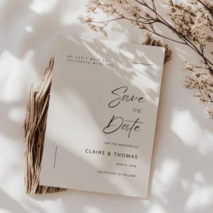 Puede incluir: Una tarjeta de invitación de boda de color crema con una elegante escritura en negro. La tarjeta dice "Save the Date" e incluye los nombres Claire & Thomas y la fecha 6 de junio de 2026. La tarjeta está sobre una base de madera con flores secas.