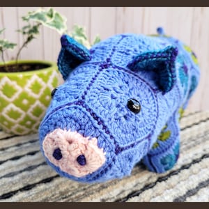 Puede incluir: Un cerdo azul de crochet con un hocico rosa y ojos negros. El cerdo está acostado sobre una alfombra rayada.