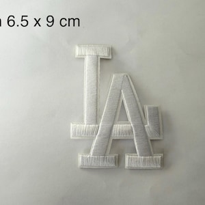 May include: White embroidered patch with the letters "LA". The patch is 6.5 cm wide and 9 cm high. The letters are in a serif font and filled with a textured, stitched pattern. The text "W/h 6.5 x 9 cm" is in the top left corner.
