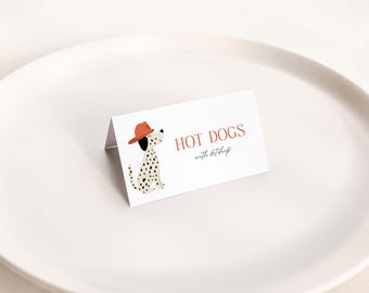 May include: A white place card with the words "HOT DOGS with ketchup" in a serif font. A cartoon Dalmatian dog wearing an orange hat is on the left. The place card is on a white plate.