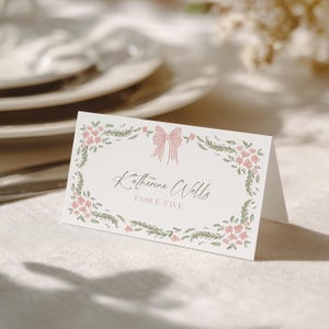 May include: A folded place card with a floral design. The card features a pink bow and floral border, with the name "Katherine Wells" and "TABLE FIVE" printed in elegant script. The card is on a white surface.