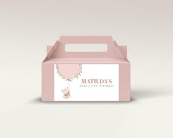 May include: A pink party favor box with a handle, featuring a graphic of a balloon, a teddy bear, and the text "MATILDA'S BEAR-Y FIRST BIRTHDAY". The box is a light pink color.