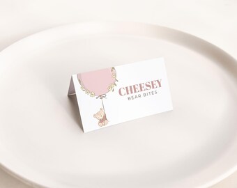 May include: A folded white card with the text "CHEESEY BEAR BITES" in a mauve color. The card features a pink balloon illustration with a small teddy bear hanging from it. The card is on a white plate.