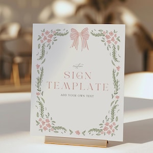 May include: A sign template with a floral design and a pink bow at the top. The sign reads "SIGN TEMPLATE" and "ADD YOUR OWN TEXT" in a soft, elegant font. The sign is displayed in a gold metal holder.