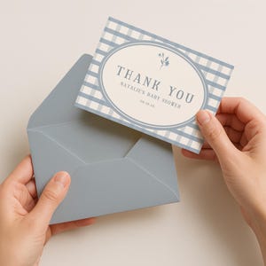 Op de afbeelding: Een bedankkaart voor een babyshower met een blauw-wit ruitpatroon en een ovale lijst. De kaart luidt "THANK YOU" en "Natalie's Baby Shower 06.16.24". De kaart wordt met een blauwe envelop vastgehouden.
