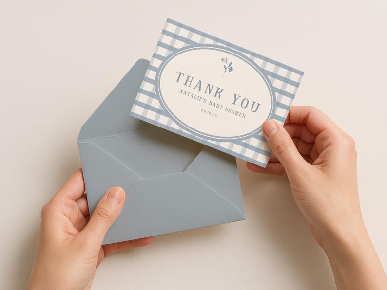 Op de afbeelding: Een bedankkaart voor een babyshower met een blauw-wit ruitpatroon en een ovale lijst. De kaart luidt "THANK YOU" en "Natalie's Baby Shower 06.16.24". De kaart wordt met een blauwe envelop vastgehouden.