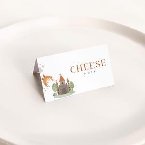Puede incluir: Una tarjeta blanca plegada con las palabras "CHEESE PIZZA" en letras marrones. La tarjeta presenta una ilustración de acuarela de un castillo, árboles y un dragón. La tarjeta está sobre un plato blanco.