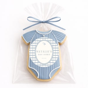 Op de afbeelding: Een baby shower koekje in de vorm van een blauwe onesie, verpakt in een doorzichtige cellofaan zak en vastgebonden met een blauw lint. De koek heeft een ovaal label met de tekst "NATALIE'S BABY SHOWER" en de datum.