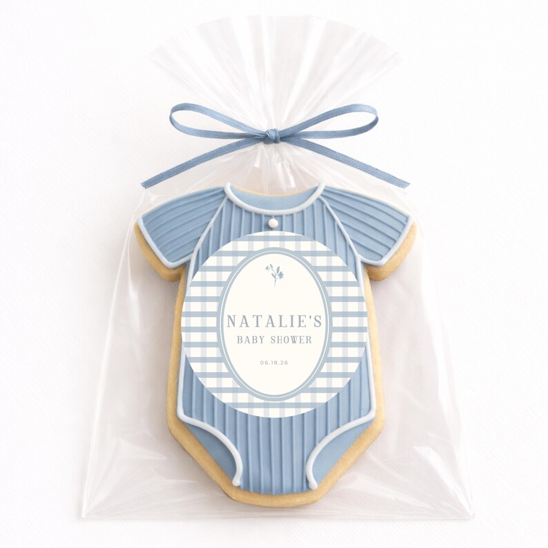 Op de afbeelding: Een baby shower koekje in de vorm van een blauwe onesie, verpakt in een doorzichtige cellofaan zak en vastgebonden met een blauw lint. De koek heeft een ovaal label met de tekst "NATALIE'S BABY SHOWER" en de datum.