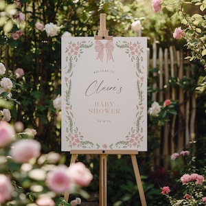 May include: A baby shower sign on a wooden easel. The sign is white with a pink bow at the top and floral border. Text reads "Welcome to Claire's Baby Shower 04.16.25". The background is a garden with pink and white roses.