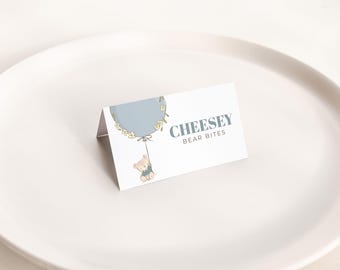 May include: A white tent card with the text "CHEESEY BEAR BITES" in gray. The card features a light blue balloon design with a teddy bear hanging from it. The card is on a white plate.