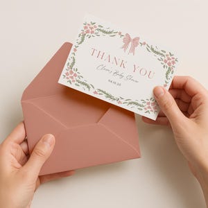 May include: A blush pink envelope holds a floral thank you card. The card features a floral wreath, a bow, and the words "THANK YOU" and "Clara's Baby Shower". The date "04.10.25" is also printed on the card.