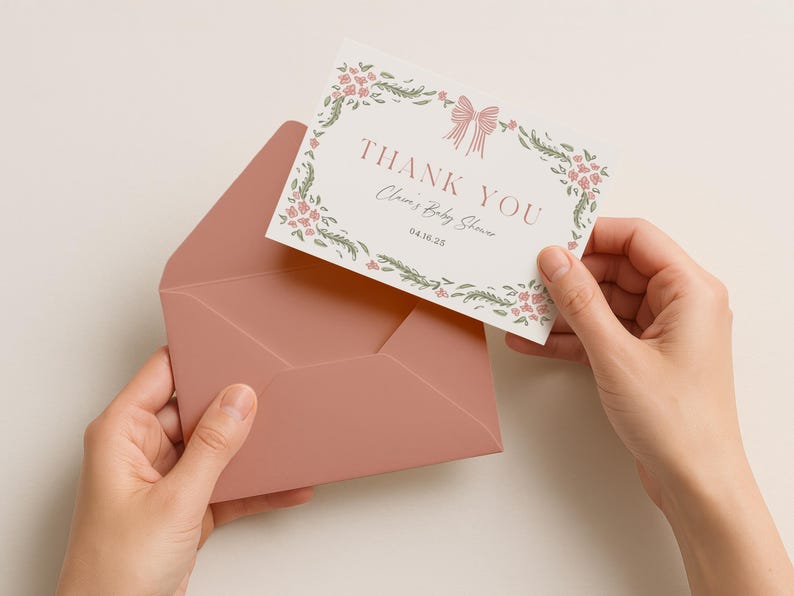 May include: A blush pink envelope holds a floral thank you card. The card features a floral wreath, a bow, and the words "THANK YOU" and "Clara's Baby Shower". The date "04.10.25" is also printed on the card.