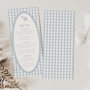 Może przedstawiać: Karta menu z niebiesko-białym wzorem w kratkę i owalną ramką z kwiatowymi akcentami. Menu zawiera przystawki, dania główne i desery. Obecna jest również pasująca karta i biała wstążka.