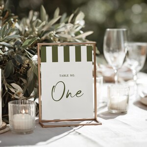 May include: A table number sign with a gold frame, featuring a white background with green and white stripes at the top. The sign reads "Table No. One" in a script font. A lit candle and greenery are in the background.