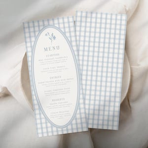 Op de afbeelding: Twee menukaarten met een lichtblauw en wit ruitpatroon. E&eacute;n kaart heeft het woord "MENU" in een ovale lijst met bloemaccenten. Het menu bevat voorgerechten, hoofdgerechten en desserts. De kaarten liggen op een wit oppervlak.