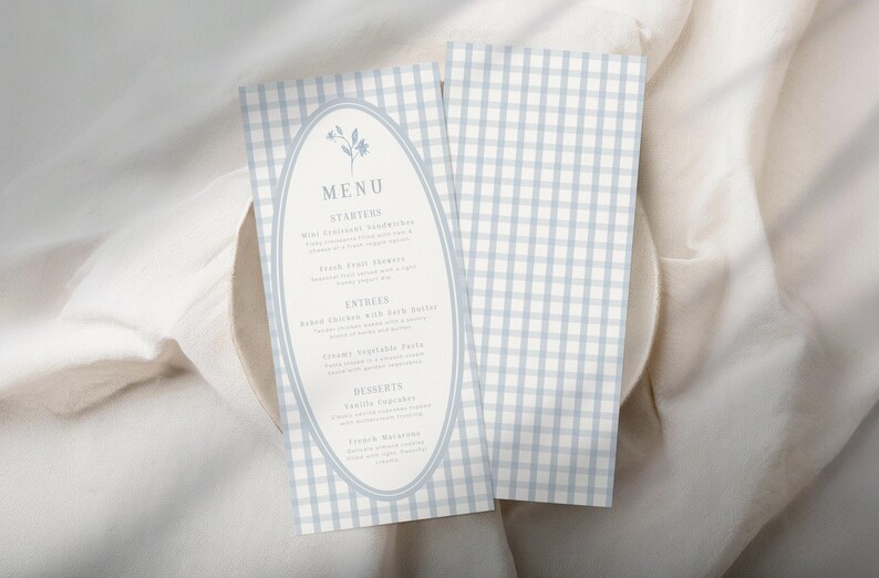 Op de afbeelding: Twee menukaarten met een lichtblauw en wit ruitpatroon. E&eacute;n kaart heeft het woord "MENU" in een ovale lijst met bloemaccenten. Het menu bevat voorgerechten, hoofdgerechten en desserts. De kaarten liggen op een wit oppervlak.