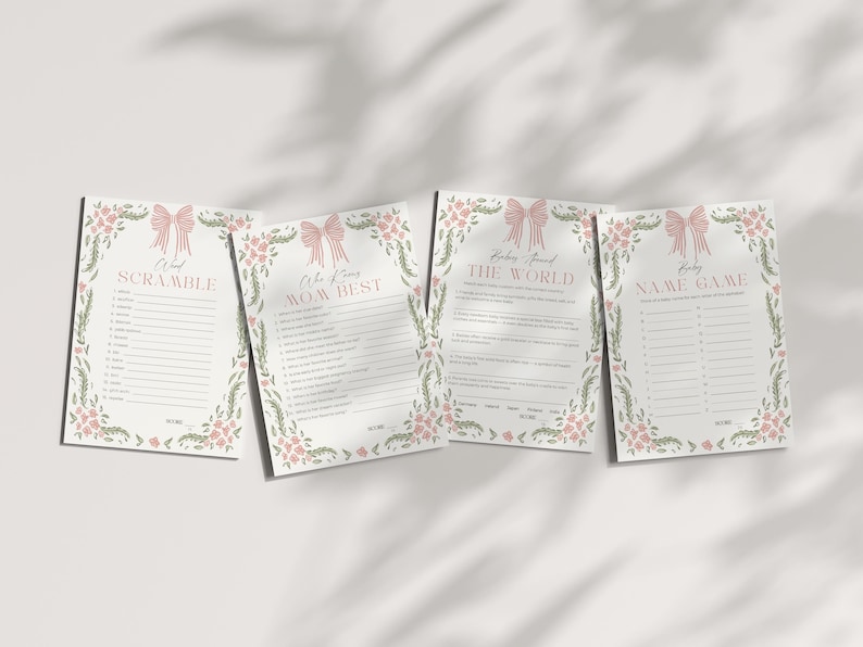 May include: Four baby shower game cards with floral and bow designs. The cards include games like "Word Scramble" and "Who Knows Mom Best". The cards are white with pink and green accents, and are arranged on a white surface.